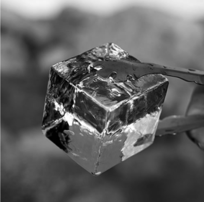 Black-and-white close-up of a rectangular ice cube held by metal tongs, with water droplets glistening on its surface against a blurred background.